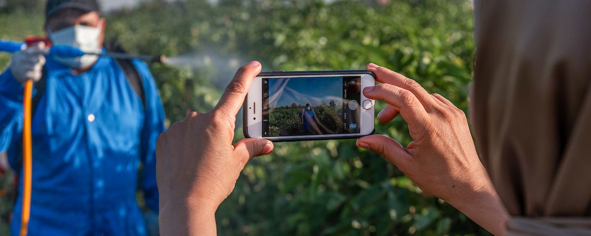 A persons hand holding a smartphone to film a farmer spraying his crops